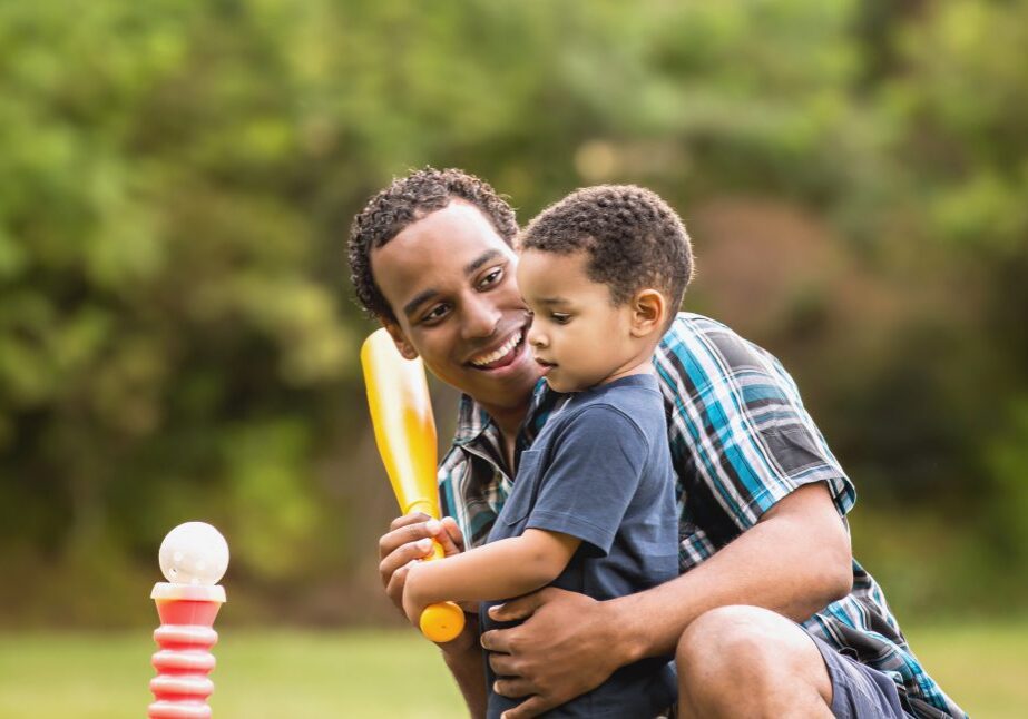 Cute African American father and son smiling in an outdoor park - lots of green background. Playing T Ball.